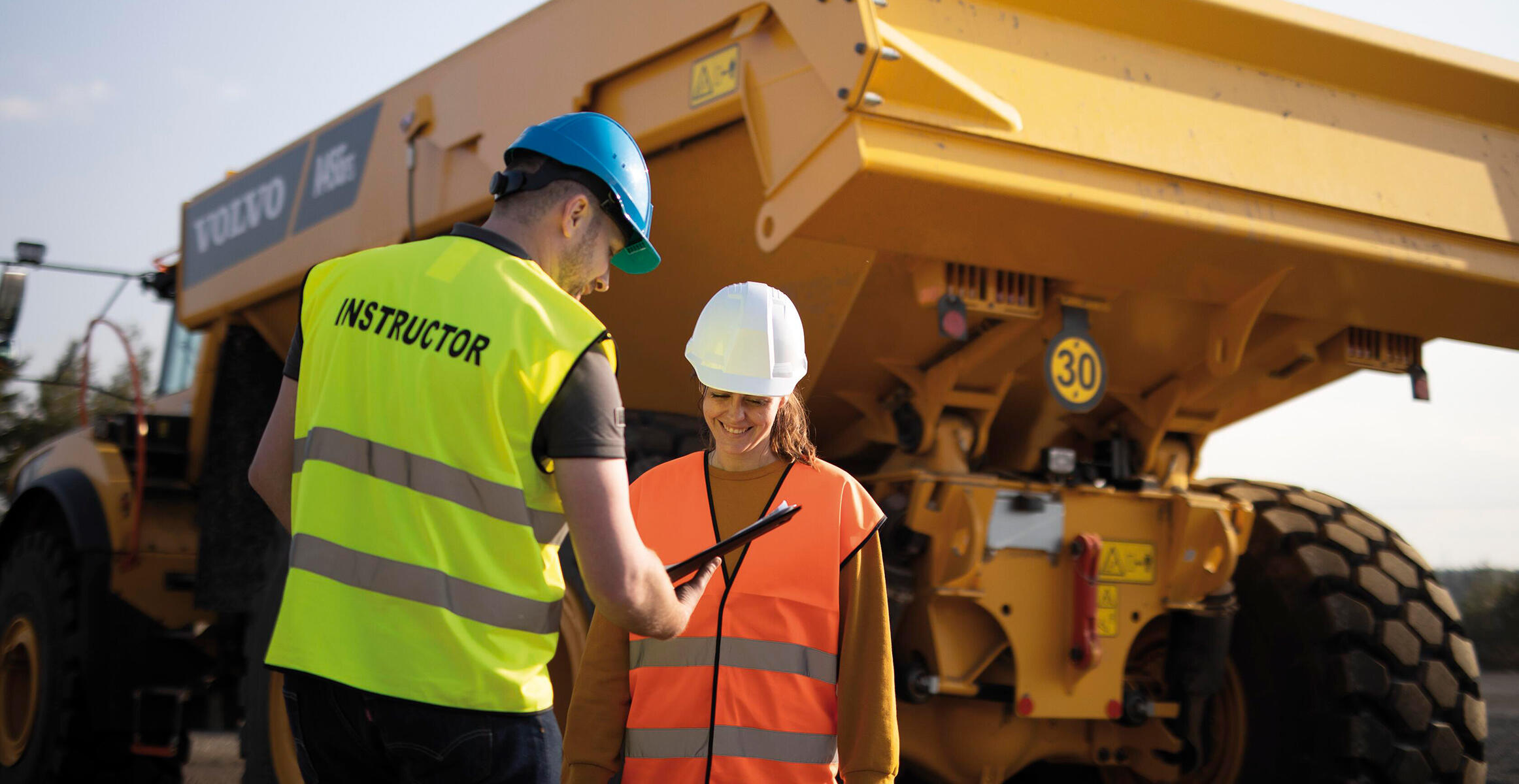 A Volvo operator training instructor shows feedback on a tablet to a smiling trainee.
