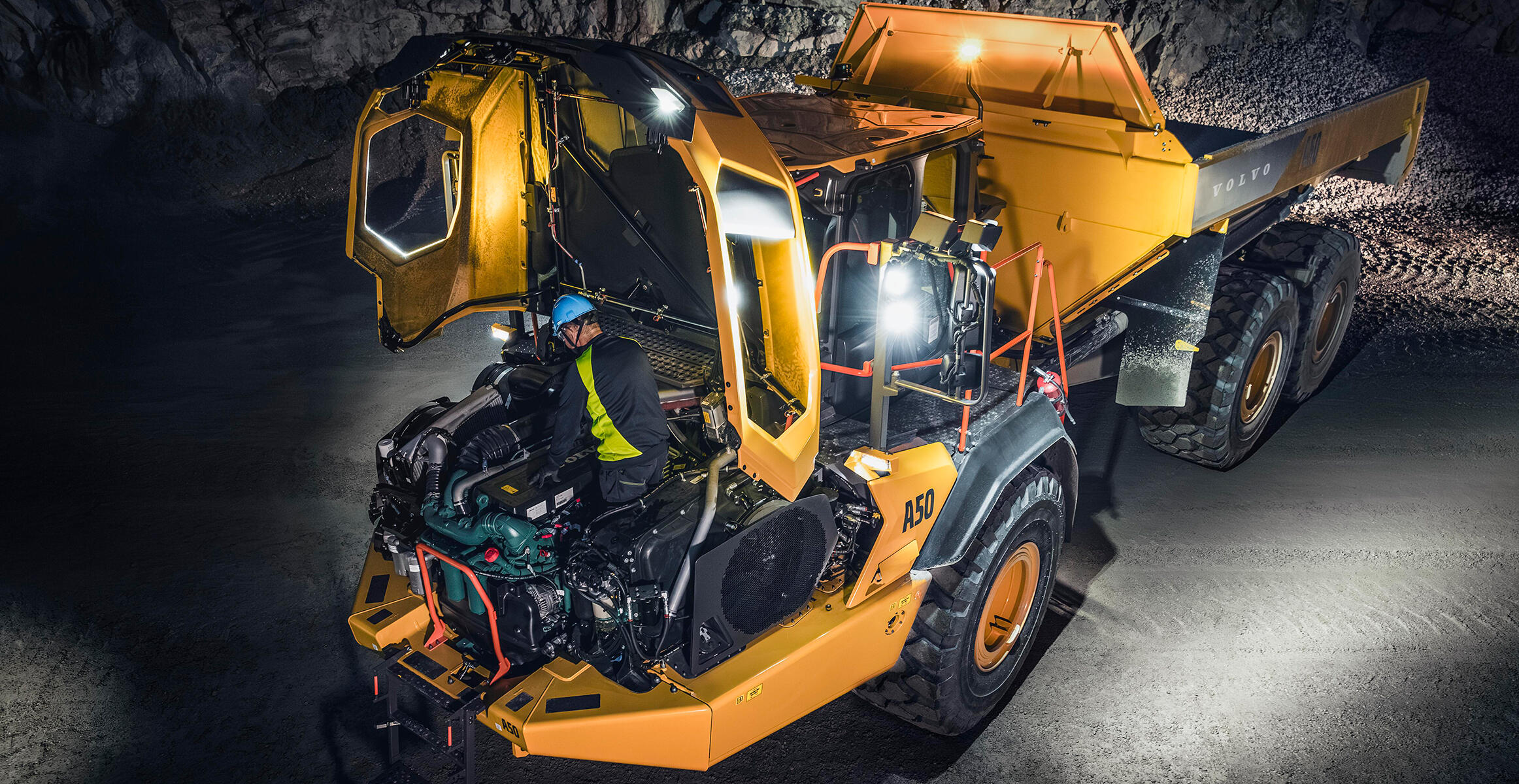 A Volvo technician is working inside the engine compartment of a Volvo Articulated Hauler at night in a quarry environment. Lights around the machine light up the area the engineer is working in.