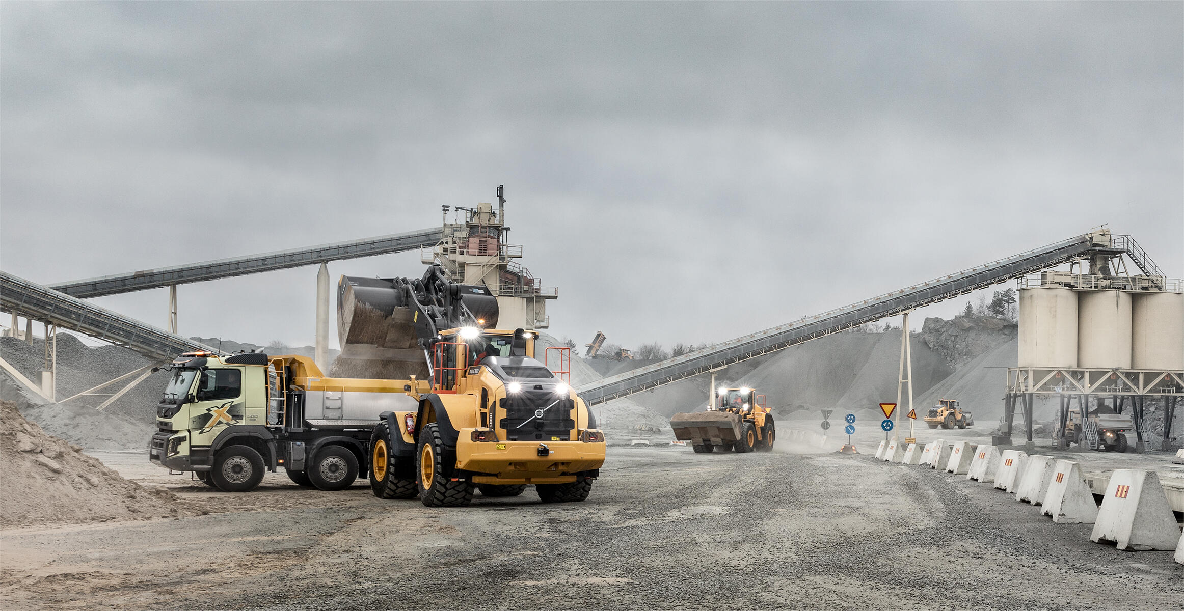 Volvo wheel loader and trucks operating at a quarry site with conveyor belts and aggregate piles