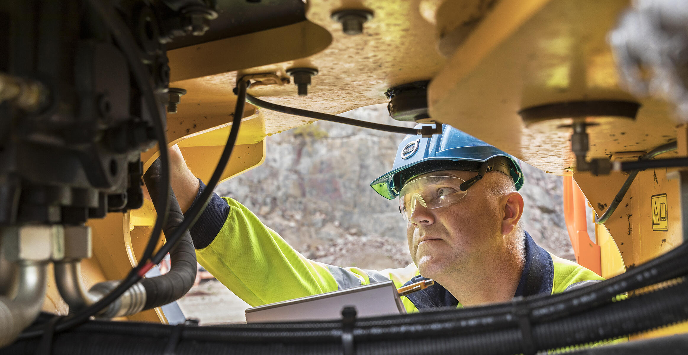Technician in safety gear performing maintenance inspection underneath Volvo construction equipment