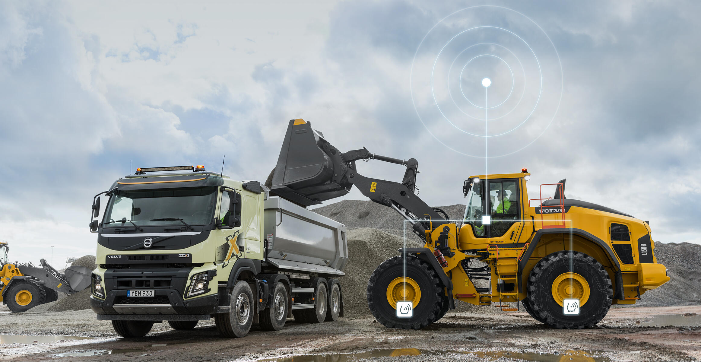 Volvo wheel loader loading material into dump truck with connectivity icon overlay at worksite